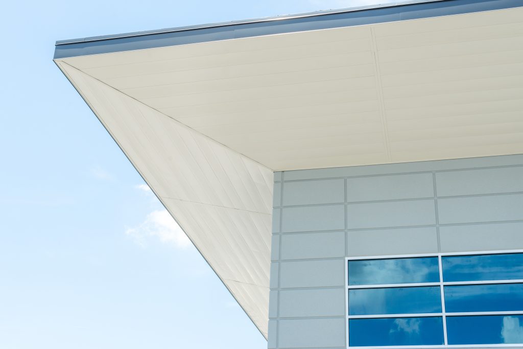 The exterior corner of a modern building with a flat roof, grey eave, white soffit, and contemporary metal aluminum composite panels. There are small blue glass windows with the blue sky reflecting.